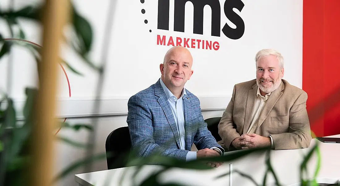 Kevin Moran and Marty Martin of IMS Marketing, a Locomotive Agency sitting in front of a board that has IMS branding on it.