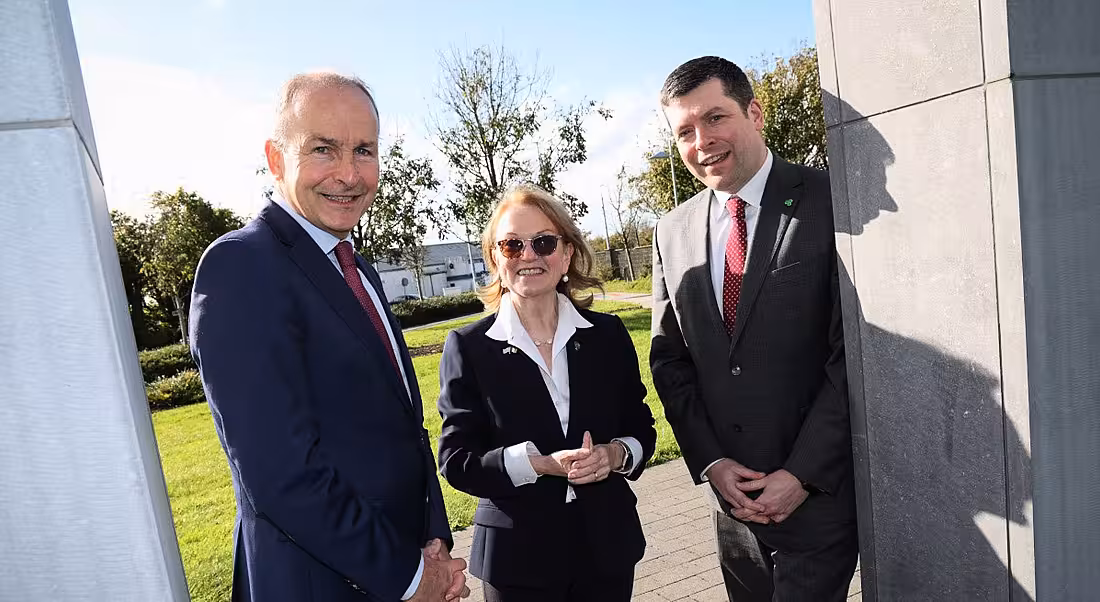 Three people standing outside on a fine day with concrete walls on either side of the image and green grass visible behind them.