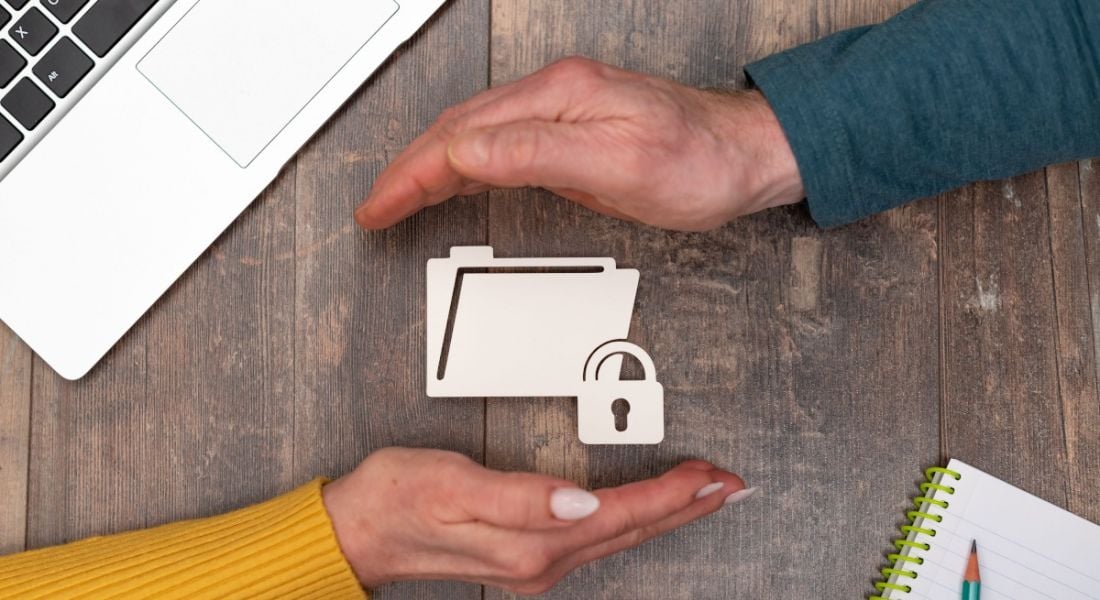 Two hands protecting a white folder icon with data. There is a laptop and a wooden table in the background.