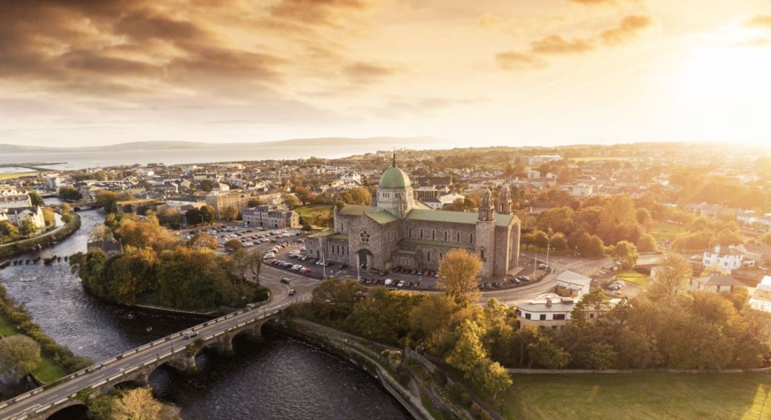 Aerial show of Galway, with a cathedral and other buildings visible and the sun shining in the background.