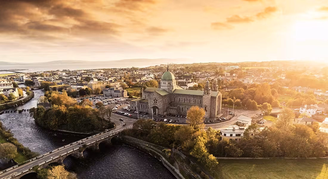 Aerial show of Galway, with a cathedral and other buildings visible and the sun shining in the background.