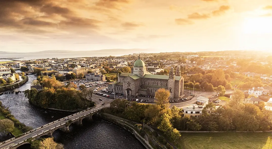 Aerial show of Galway, with a cathedral and other buildings visible and the sun shining in the background.