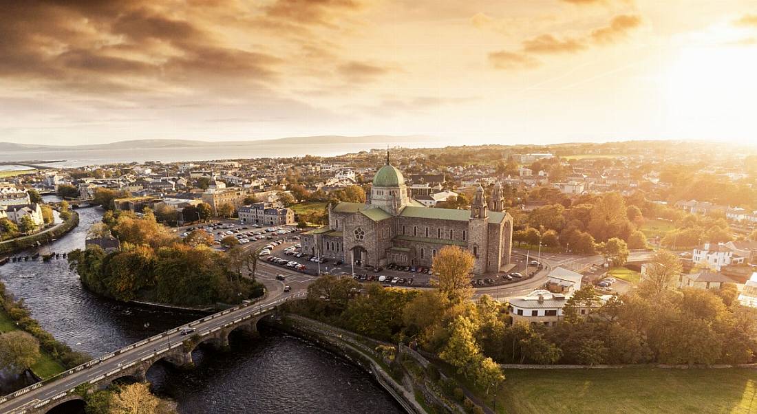 Aerial show of Galway, with a cathedral and other buildings visible and the sun shining in the background.