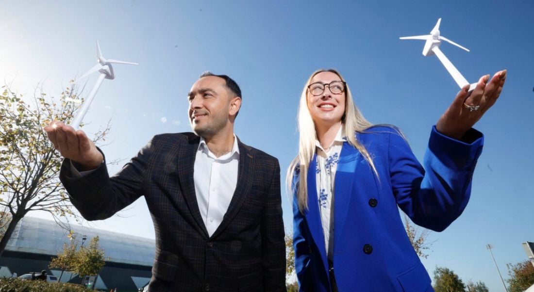 Two people posing near wind turbines with a bright blue sky behind them.