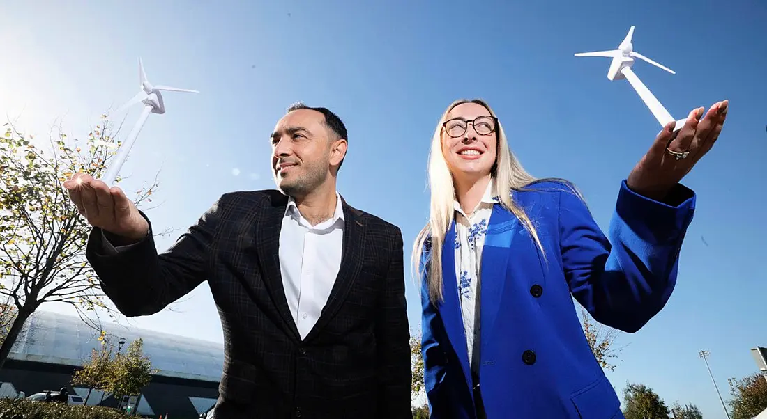 Two people posing near wind turbines with a bright blue sky behind them.