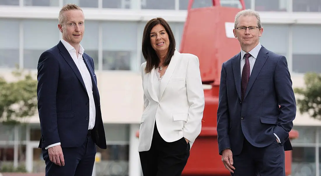 Three people standing outside a building with a glass front and a red structure in front of it.