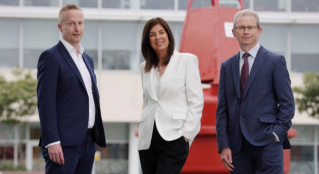 Three people standing outside a building with a glass front and a red structure in front of it.