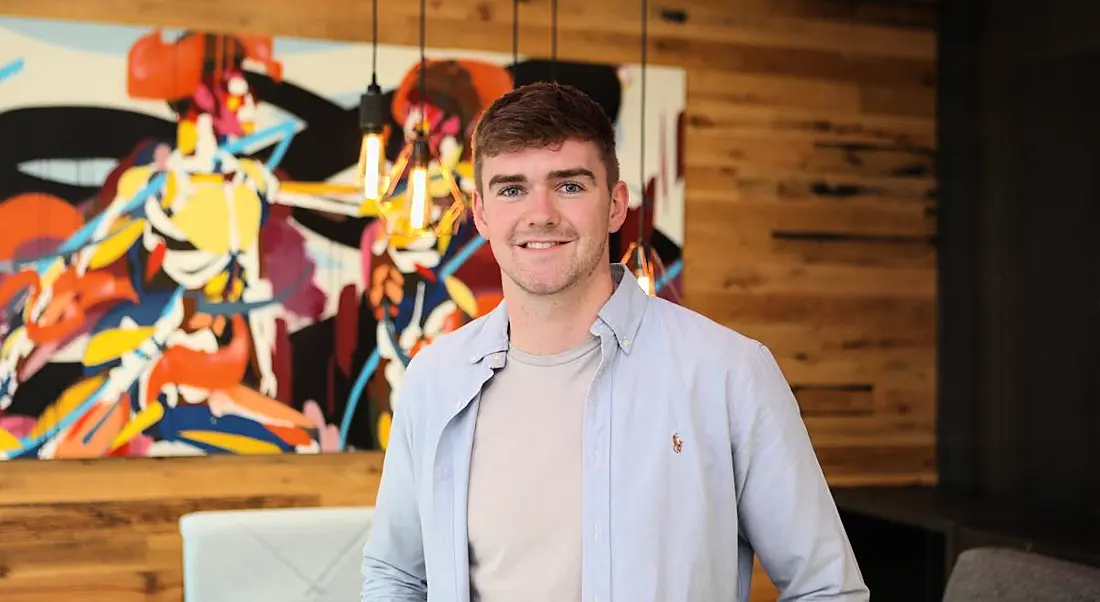 A man smiles at the camera in front of a wood panel wall. There is a colourful abstract painting on the wall. The man is Dara Bell, a technology consultant at Accenture.