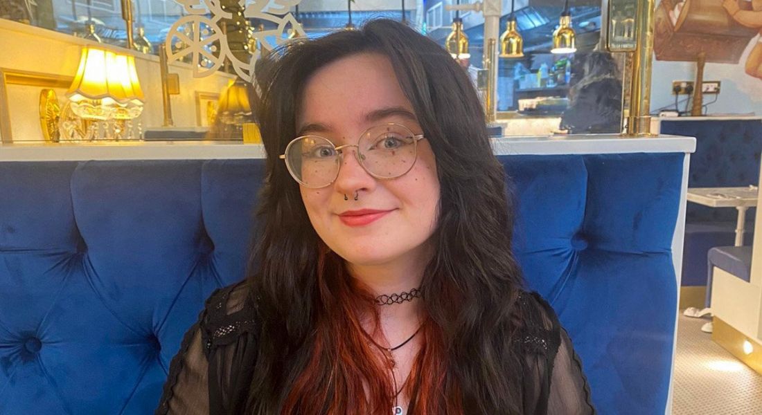 A woman with long dark hair with the ends dyed red smiles at the camera while sitting in a restaurant booth on a blue seat. She is Emma Louise Bonner, a software engineering intern at Liberty IT.