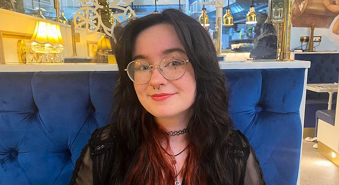 A woman with long dark hair with the ends dyed red smiles at the camera while sitting in a restaurant booth on a blue seat. She is Emma Louise Bonner, a software engineering intern at Liberty IT.