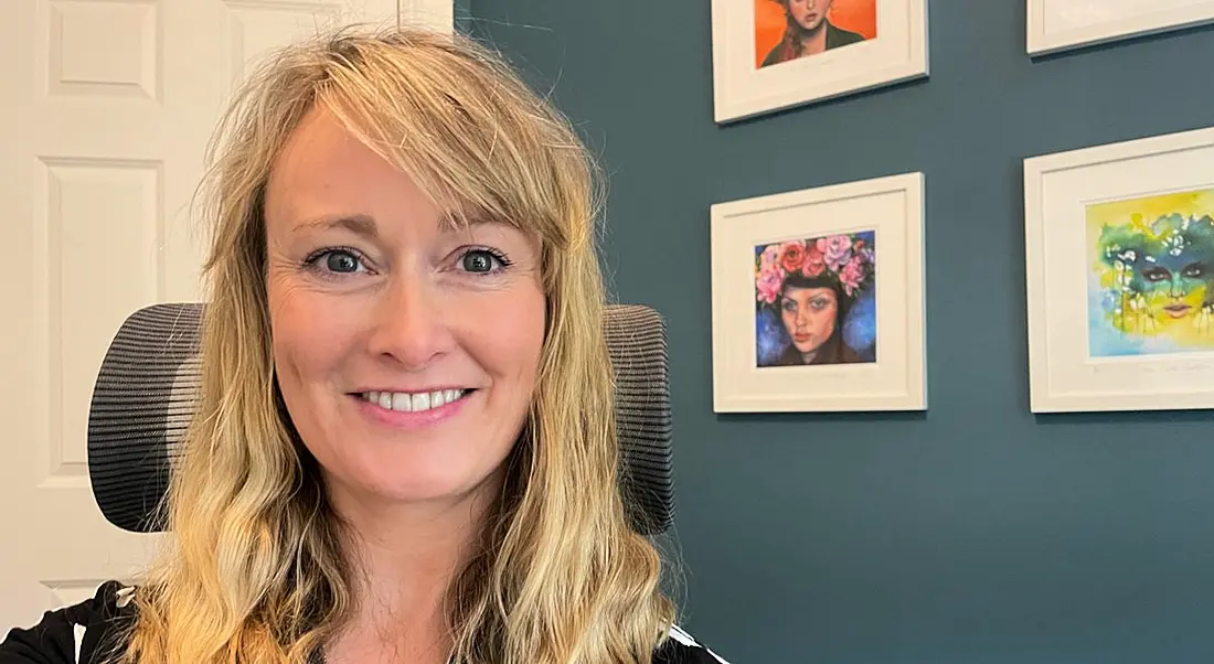 Stephanie Sheehan sitting in an office chair facing the camera with paintings visible on a wall behind her.