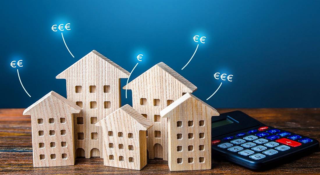 An image of a group of model houses sitting next to a calculator on a table. There are blue euro signs hovering outside each of the houses with a line connecting them to the houses.