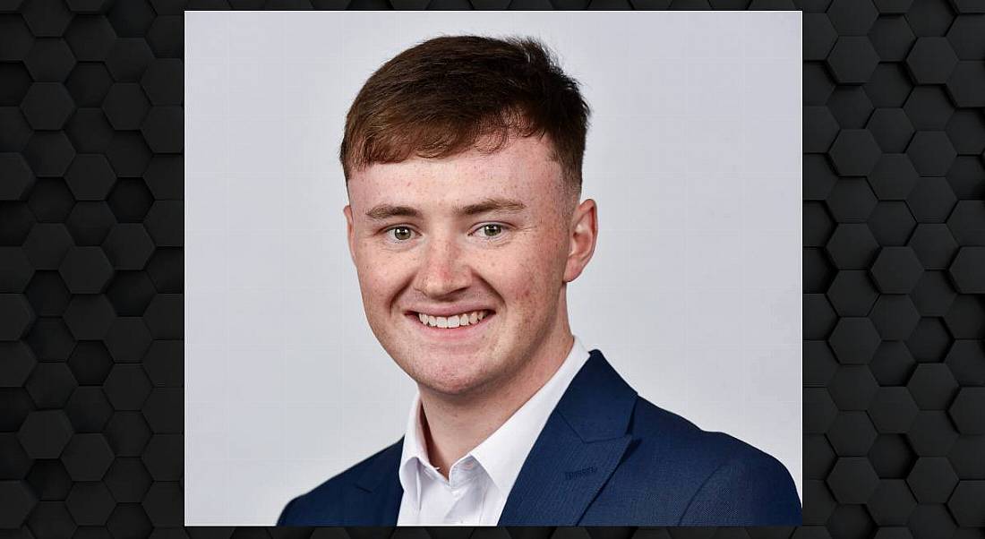 A man wearing a navy suit with a white shirt and no tie smiles at the camera in front of a grey background. He is Neil Toner, an analyst working at Aon.