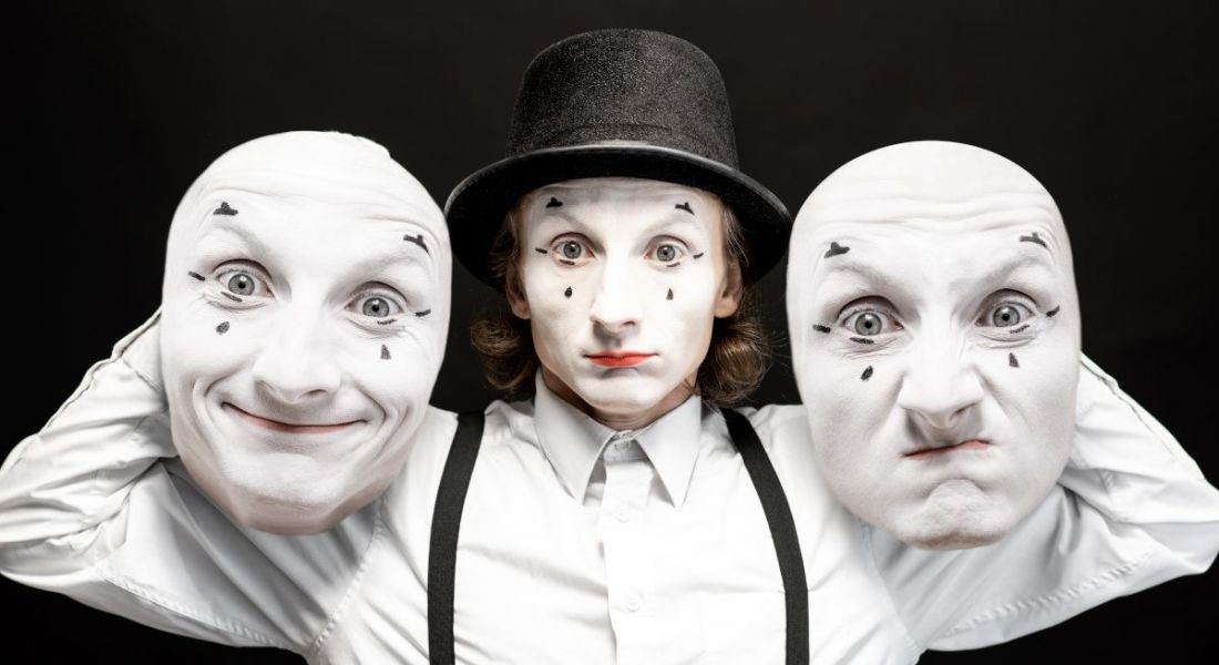 A mime dressed in white with white face paint holding two white mime masks either side of their face against a black background.