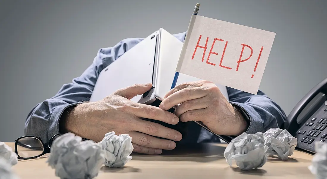 Worker with his head down on his desk, overworked and frazzled. There are scrunched up pieces of paper on the desk and he is holding a flag that reads 'Help!'