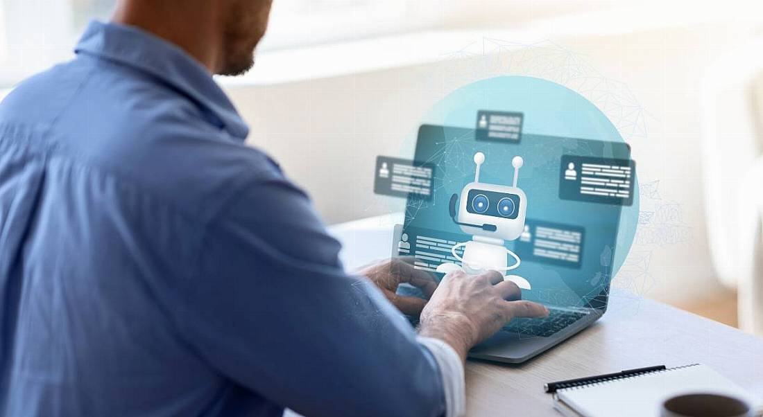 A man typing on a laptop at a desk, with an image of a virtual chatbot in front of the laptop screen. Used to represent the concept of Generative AI.