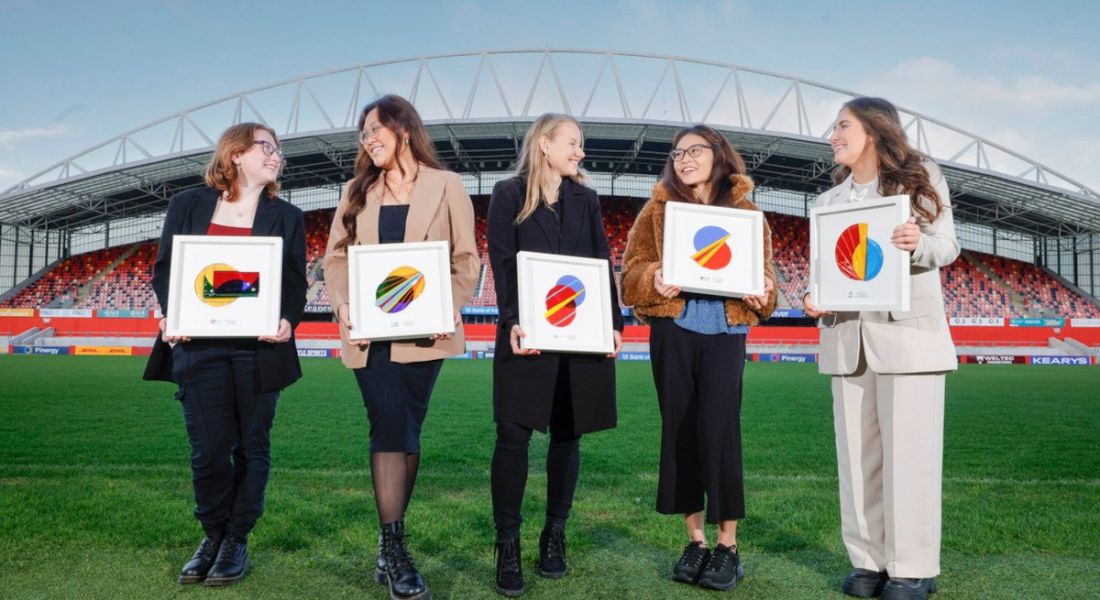 Five young women standing in a row at Thomond Park stadium holding art pieces.