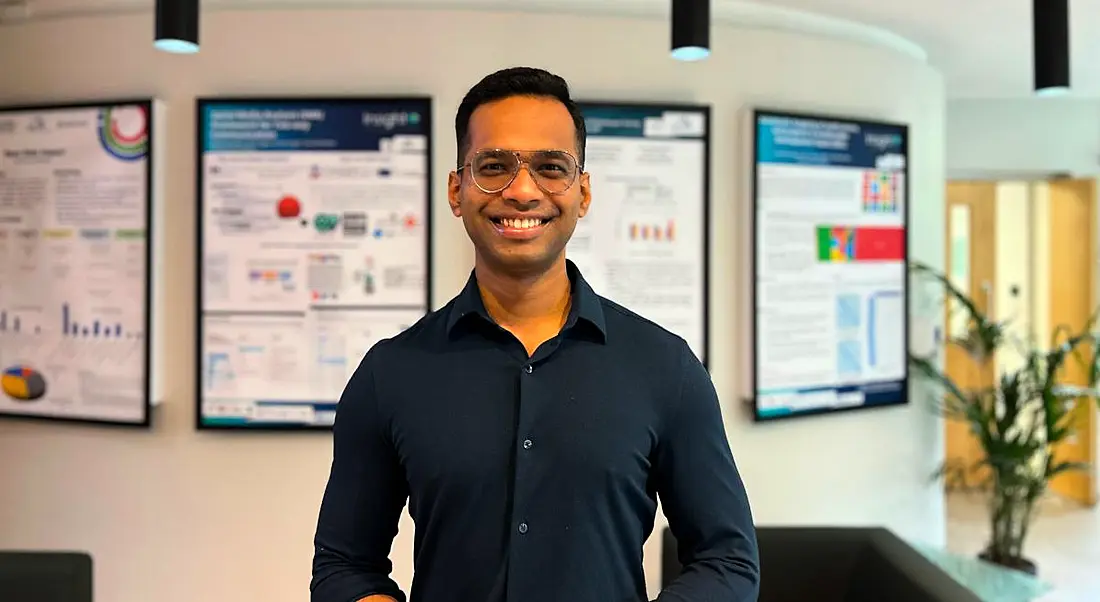 A man wearing a navy-coloured shirt and glasses smiles at the camera in front of a wall that holds various framed charts. He is Rajdeep Sarkar, a research scientist at Yahoo.