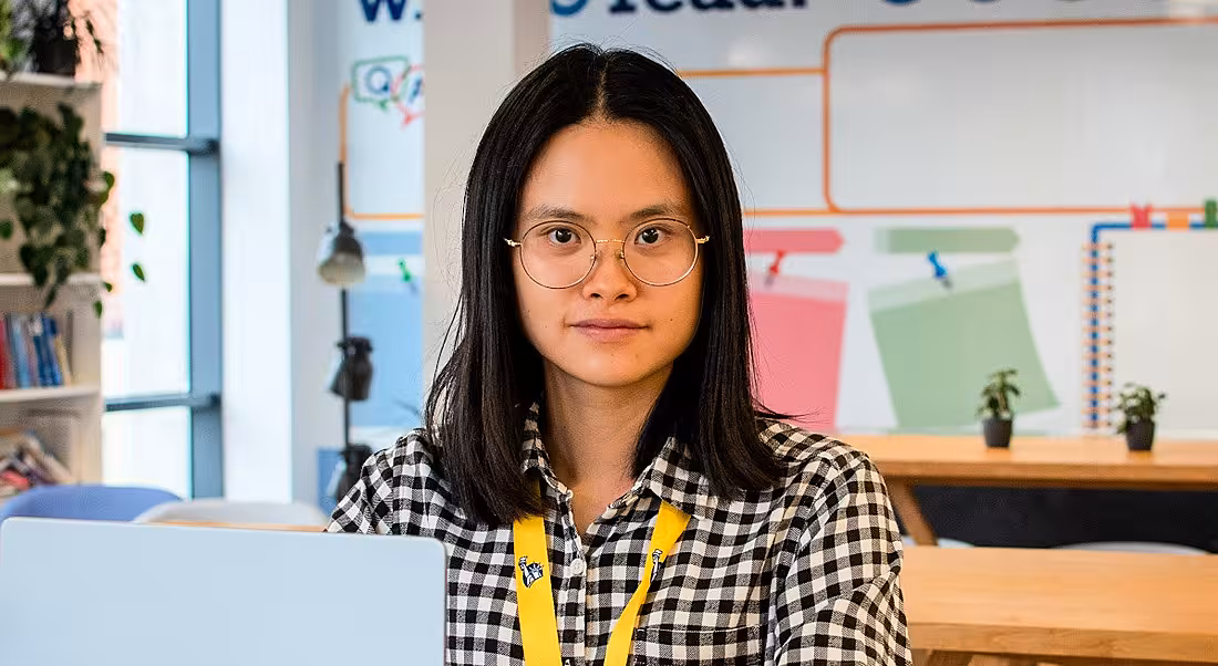 A woman wearing glasses and a chequered shirt smiles at the camera from behind a laptop. She is Wanqi Chen, an associate software engineer at Liberty IT.