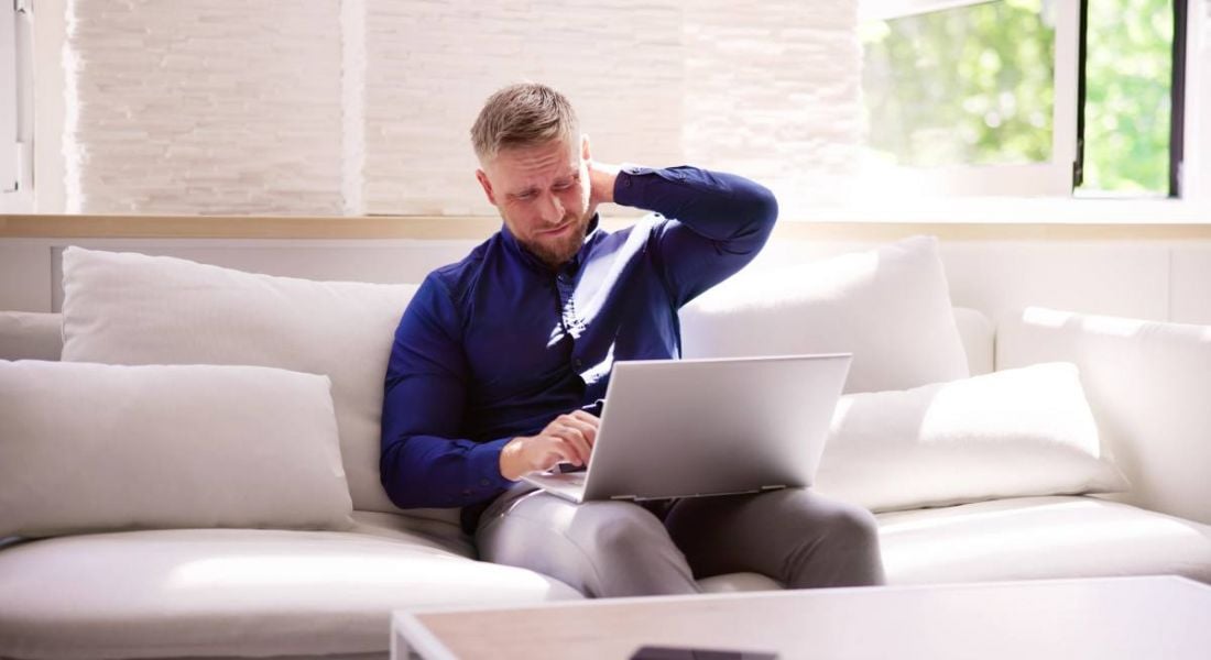 A man working on a laptop while sitting on a couch holds his neck with his left arm out of pain.