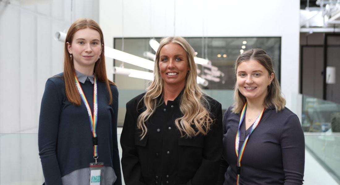 Three women smile while standing next to each other in an office building hallway. They are all involved in Amgen's biotech graduate programme.