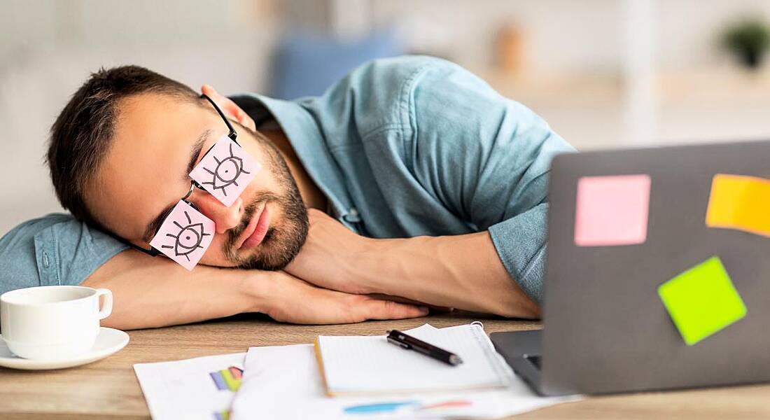 A man napping at a messy desk beside a laptop. He has pink Post-it notes with eyes drawn on covering his own eyes, showing employee disengagement.