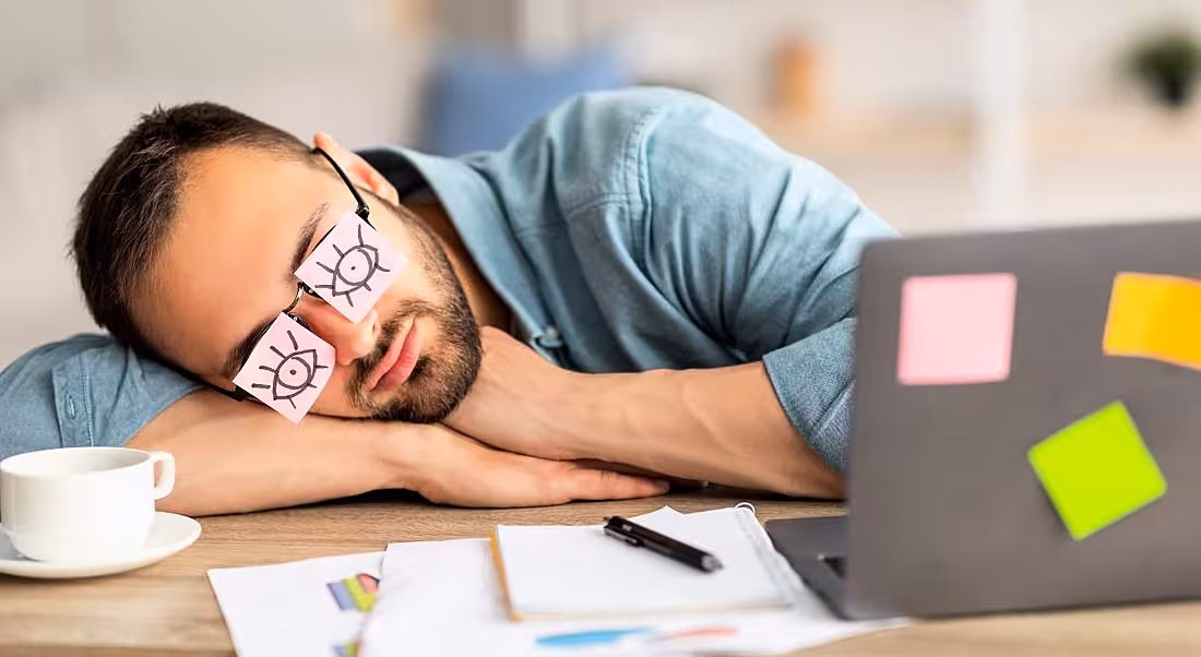 A man napping at a messy desk beside a laptop. He has pink Post-it notes with eyes drawn on covering his own eyes, showing employee disengagement.