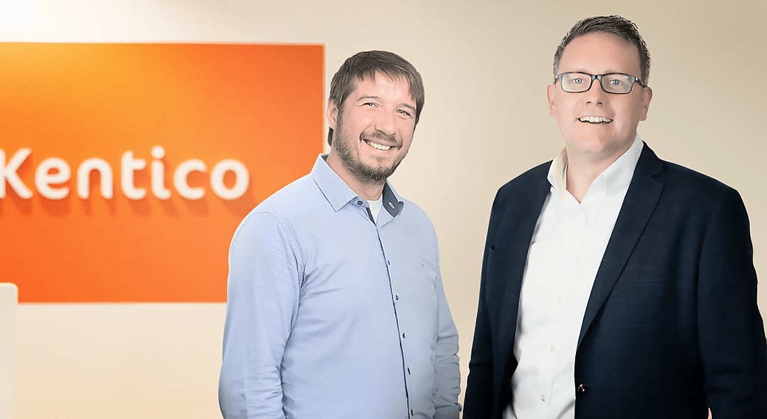 Two men standing together in front of a wall that has the Kentico logo on it. They are part of Kentico and Granite Digital.