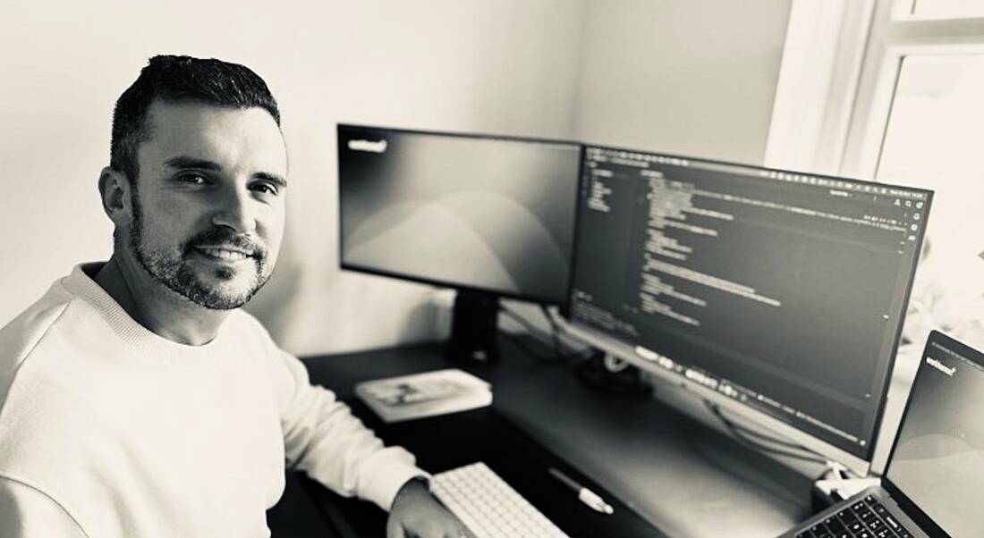 A man wearing a light-coloured jumper sits at a work desk and smiles to the camera. On his desk are two computer monitors, with one of the screens displaying lines of code. He is Keith Tuffy, a principal engineer at Workhuman.