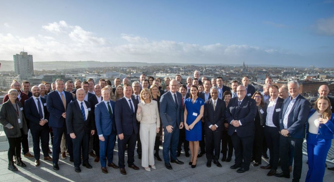 A group shot of Qualcomm team with Simon Coveney and Mary Buckley on the roof of the office with a view of Cork city behind them and a clear blue sky.
