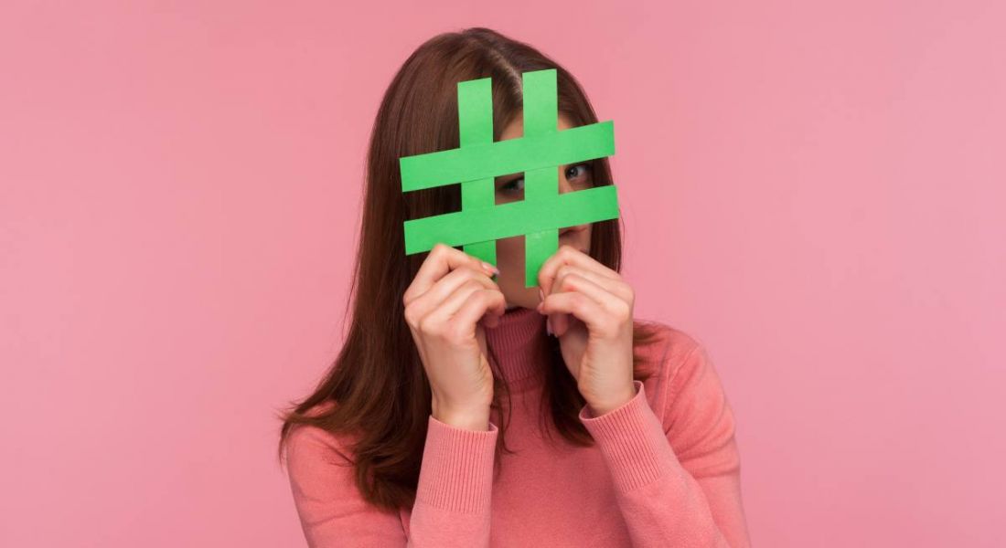Young woman holding a green hashtag in front of her face. The background behind her is bright pink.