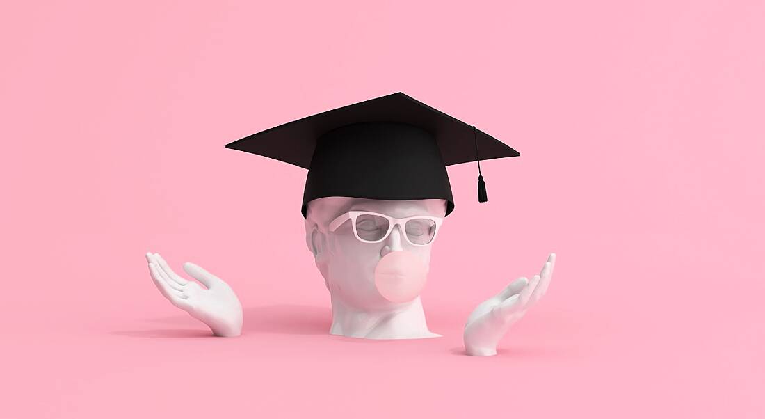 A statue of a head wearing a graduate cap and glasses blowing bubblegum on a pink background.