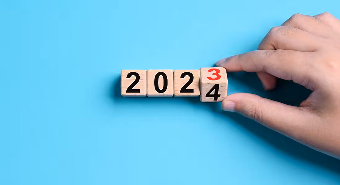Person's hand turning a cube on a calendar from 2023 to 2024. Blue background.