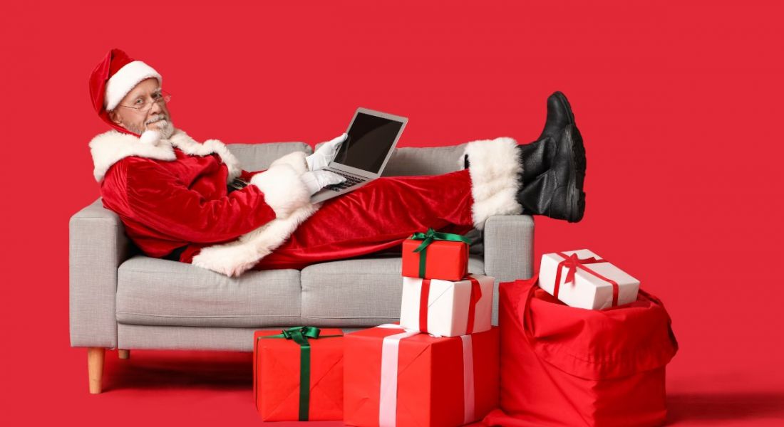 A man who looks like Santa Claus lying on a sofa with a laptop and presents on the floor beside him. The background is red and he is wearing a Santa outfit.
