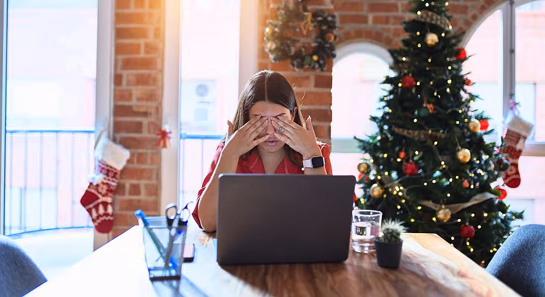 A woman rubs her eyes in front of laptop, symbolising burnout. Behind her, the office is decked out in Christmas decorations and a tree.