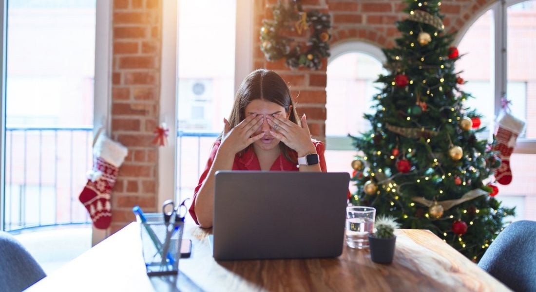 A woman rubs her eyes in front of laptop, symbolising burnout. Behind her, the office is decked out in Christmas decorations and a tree.