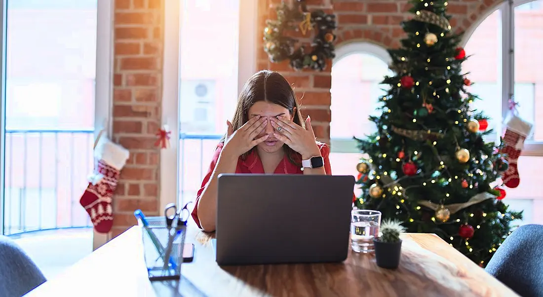 A woman rubs her eyes in front of laptop, symbolising burnout. Behind her, the office is decked out in Christmas decorations and a tree.