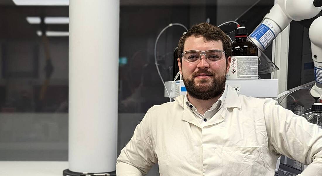 A man wearing a white lab coat and goggles smiles at the camera while resting his arm on lab equipment. He is Vincent O'Brien, an analytical chemist at Henkel.