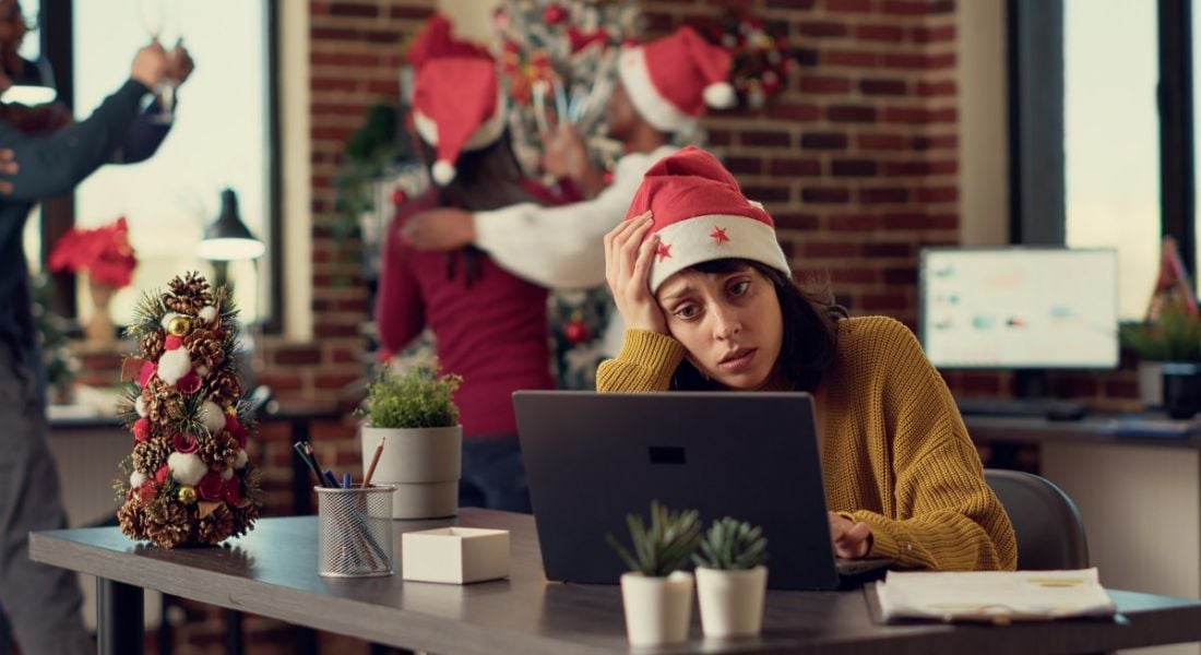 A stressed woman wearing a Santa hat sits at a desk with a laptop in front of her while she holds her head with one of her hands. There are people behind her also wearing Santa hats and celebrating.