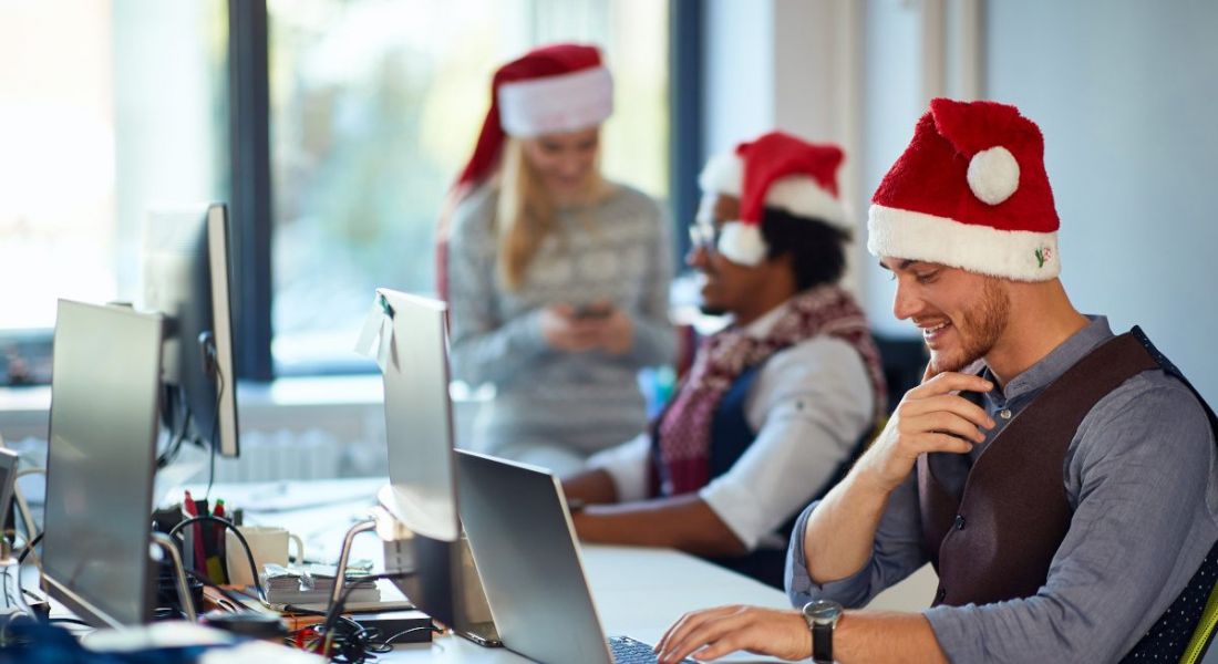 Three office workers wear santa hats while sitting at their computers in front of a big window.