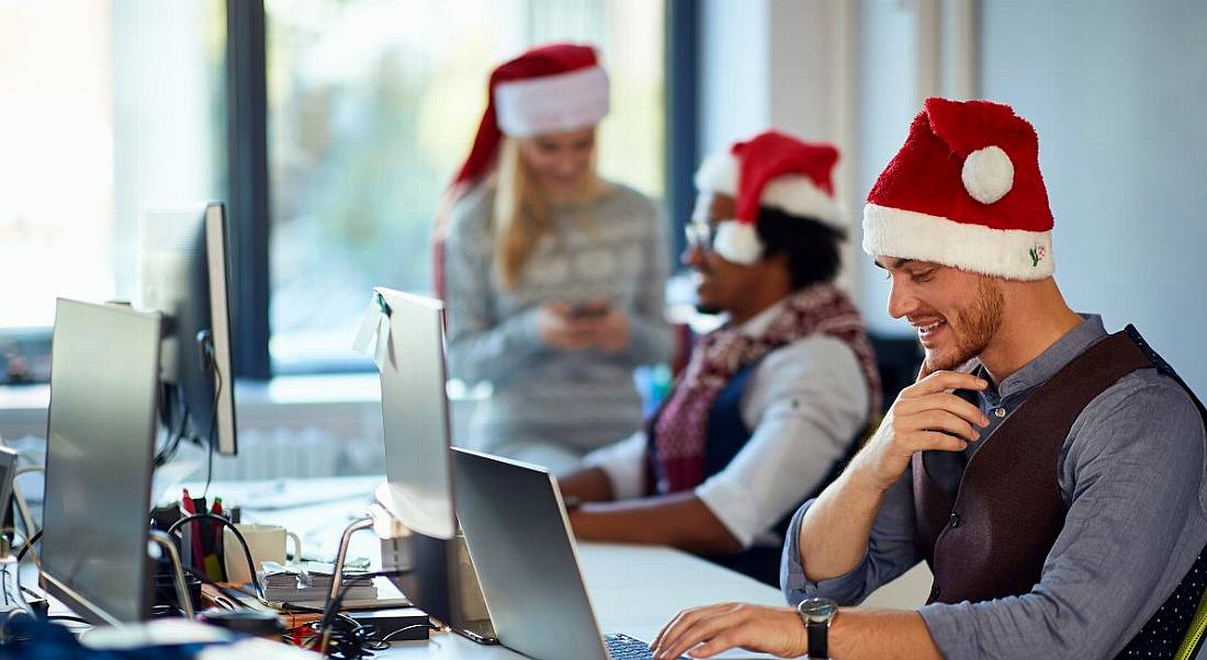 Three office workers wear santa hats while sitting at their computers in front of a big window.