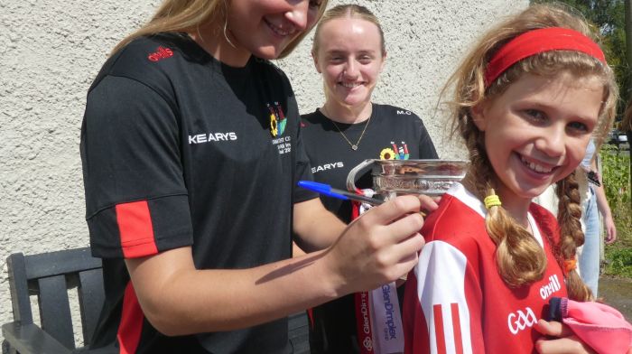 Éabha O'Leary getting her jersey signed by Cork senior camogie player Ciara O'Sullivan, with Millie Condon and the O'Duffy Cup looking on, at the opening of the sunflower field.